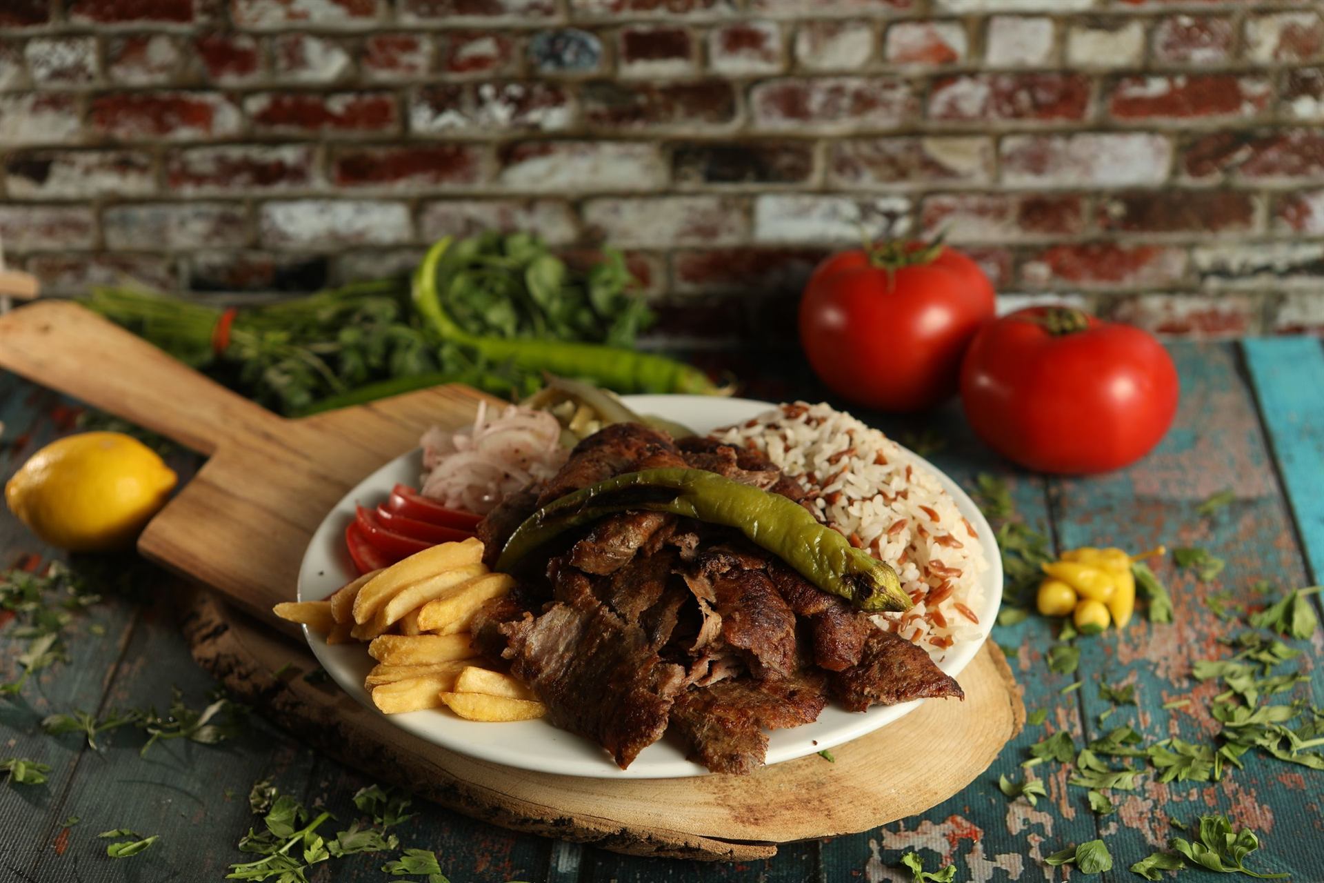 Een schotel d&ouml;ner met friet, rijst, tomaat en een gegrilde peper op een houten plank.
